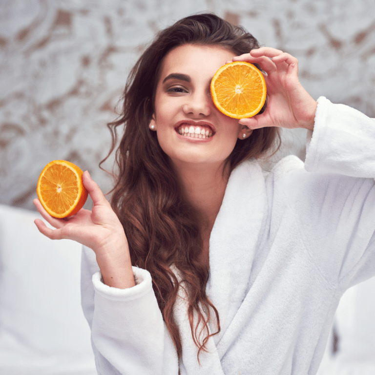 Mujer morena de frente con un albornoz blanco con media naranja en cada mano y una de ellas cubriéndose el ojo izquierdo, dando importancia a la vitamina C que aporta la naranja.